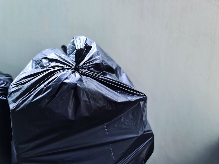 A large green waste collection bin filled with an assortment of discarded cardboard boxes, some flattened and others in their original three-dimensional form, with visible flaps and labels. The bin ap