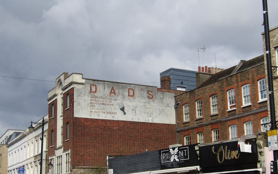 The image shows a row of buildings on a street, with a prominent weathered brick wall in the foreground displaying a faded painted sign that reads 'DAD'S' in large, red letters, along with smaller, pa
